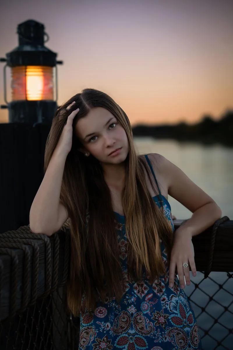Senior portrait of a young woman seated beside a glowing lantern at dusk, with a lake in the background