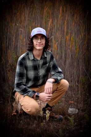 A person wearing a plaid shirt, beige pants, and a cap is crouching in a field with tall grass.