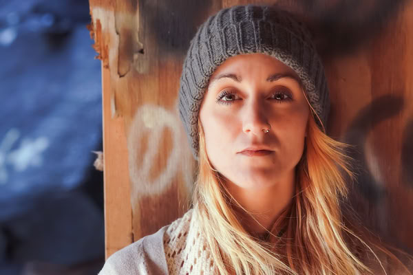 A person with long blonde hair wearing a gray knit beanie and a scarf, standing in front of a wooden surface with graffiti.