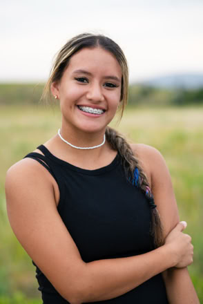 A person with long hair in a braid, wearing a black sleeveless top and a necklace, smiling in an outdoor setting with a blurred grassy background.