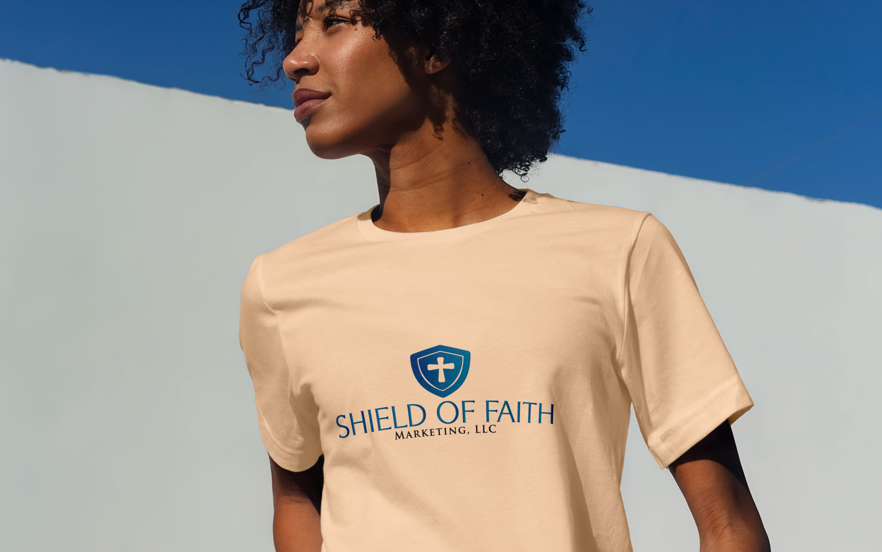 Person with curly hair wearing a beige t-shirt with a shield logo and text, standing against a blue and white background.
