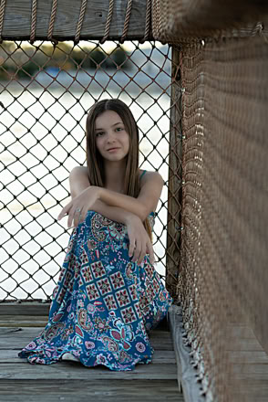A person with long hair sitting on a wooden surface, wearing a blue patterned dress, with a chain-link fence in the background.